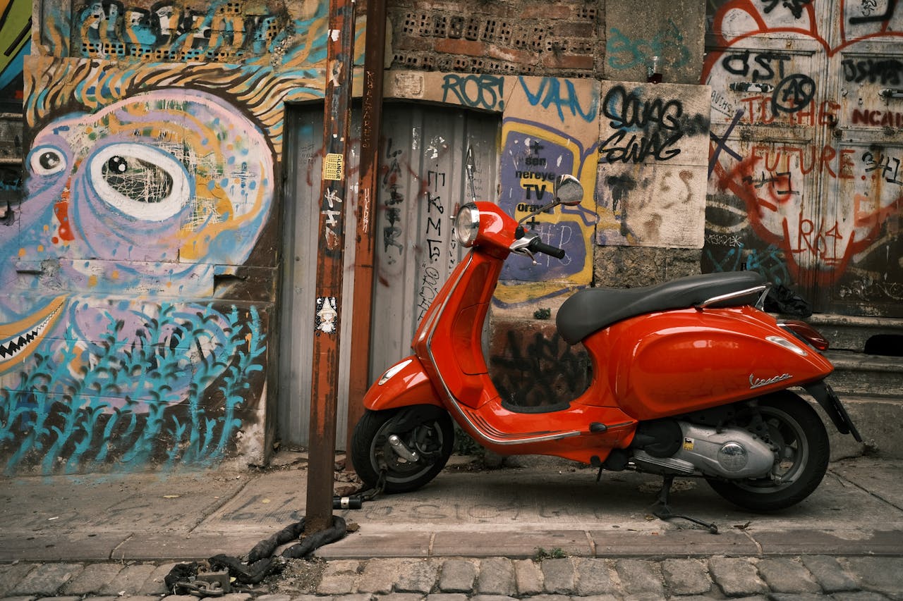gallery-1 Red Vespa parked on an urban sidewalk, with colorful graffiti background.